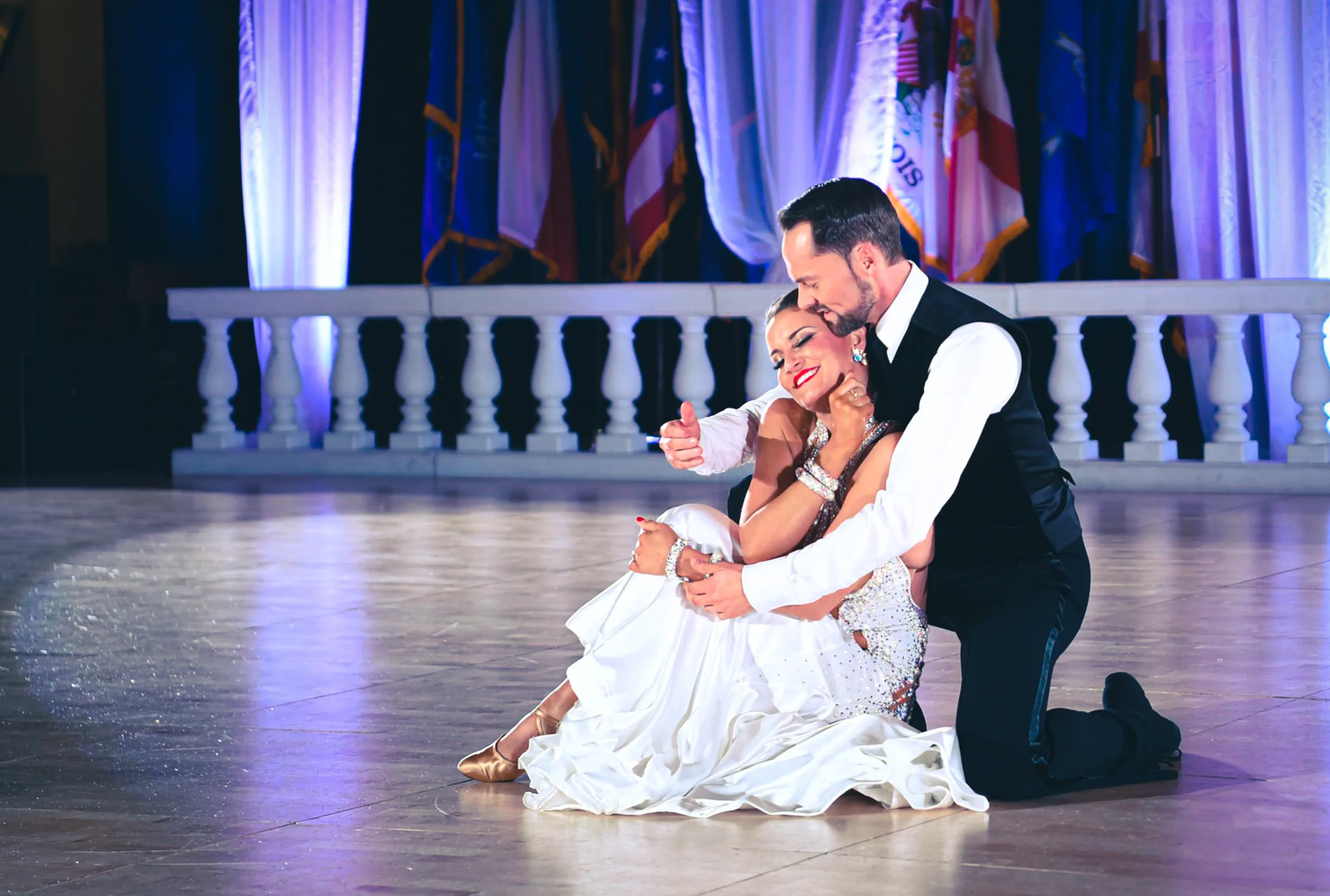 Travis kneels with his arm swept wide as Jaimee arches beside him before a stage balustrade